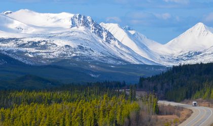 Ein Campervan fährt auf dem Alaska Highway im Yukon, Kanada, durch eine weite, bewaldete Landschaft, umgeben von Bergen unter einem bewölkten Himmel.