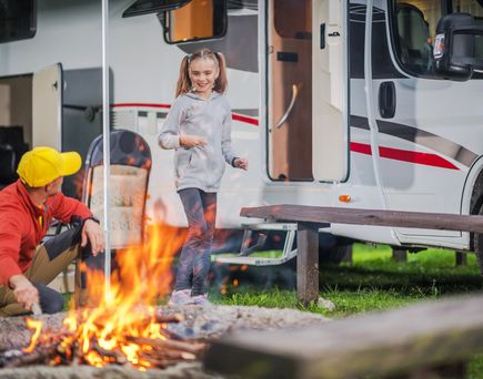 Eine Familie sitzt gemütlich um ein Lagerfeuer vor ihrem Wohnmobil, Kinder spielen.