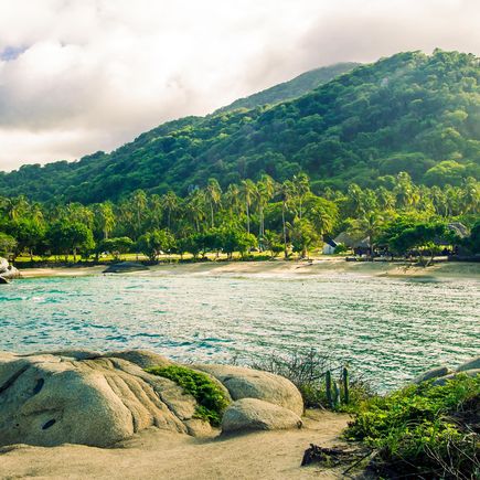 Blick auf eine idyllische Bucht im Tayrona-Nationalpark, Kolumbien, mit einem weißen Sandstrand, türkisfarbenem Wasser und tropischer Vegetation unter blauem Himmel.
