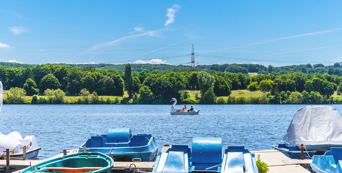 Tretboote liegen an einem Steg am Kemnader See, während im Hintergrund ein Schwanen-Tretboot auf dem Wasser fährt und grüne Bäume das Ufer säumen.