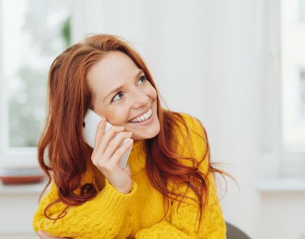 Eine rothaarige Frau in einem gelben Pullover lächelt, während sie ein Smartphone an ihr Ohr hält, offenbar im Gespräch.