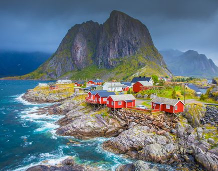 Ein idyllisches Dorf auf den Lofoten-Inseln in Norwegen, mit charmanten roten Häusern, die sich an die Küste schmiegen, umgeben von majestätischen Bergen und dem ruhigen blauen Meer.