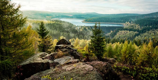 Eine atemberaubende Landschaftsaufnahme der Talsperre Fláje im Erzgebirge, gesehen von zerklüfteten Felsen im Vordergrund, umgeben von einem dichten Wald.