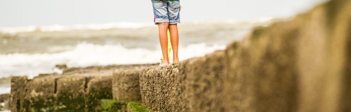 Ein Kind mit grüner Jacke und kurzer Hose spaziert auf einem Steinsteg am Strand.