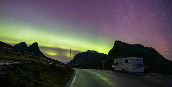 Eine spektakuläre Aufnahme der Trollstigen-Straße in Norwegen bei Nacht, die die kurvenreiche Bergstraße beleuchtet, während die Nordlichter am Himmel tanzen und die Landschaft in magisches Licht tauchen.