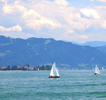 Ein Panoramablick auf den Bodensee, umgeben von malerischen Städten und grünen Hügeln, mit Booten auf dem Wasser und einem klaren blauen Himmel.
