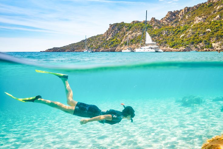 Eine Person schnorchelt in klarem, türkisfarbenem Wasser, während die Sonne die Unterwasserwelt beleuchtet. Felsformationen sind am Rand der Bucht zu sehen.