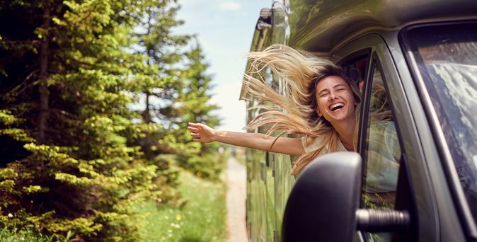 Eine fröhliche junge Frau mit wehendem blondem Haar lehnt sich lachend aus dem Beifahrerfenster eines fahrenden Wohnmobils auf einer von Bäumen gesäumten Straße.