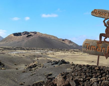 Lanzarote. Blick in der Dürre auf das Holz-Hinweisschild des Timanfaya Nationalparks. Einige Berge im Hintergrund, blauer Himmel.