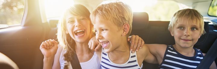 Drei fröhliche Kinder sitzen angeschnallt auf der Rückbank eines Autos und lachen in die Kamera bei Sonnenlicht.