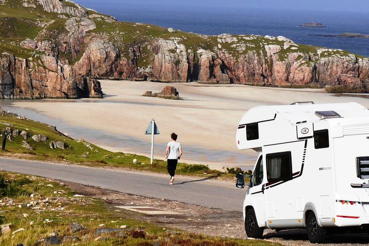 Ein Camper steht an einer Straße nähe einem Strand mit Felsen und ein Mann läuft in Richtung Strand weg vom Camper