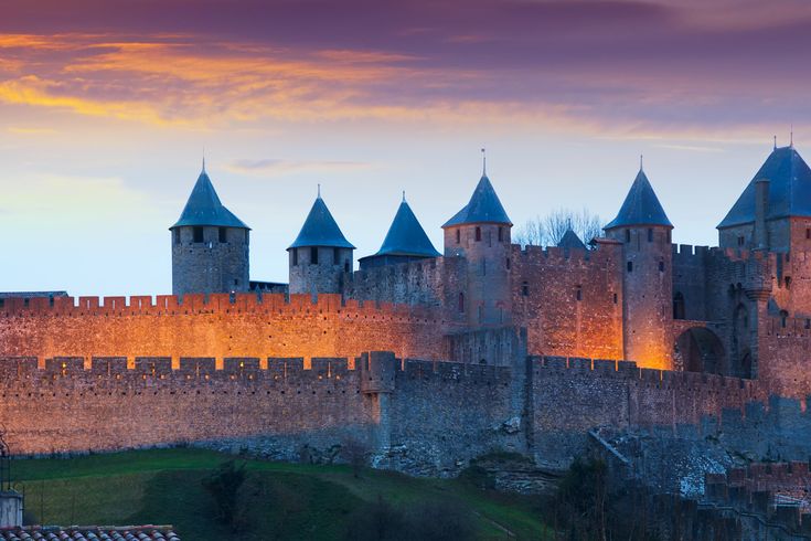 Eine imposante historische Burg oder Festung mit hohen Steinmauern und Türmen, beleuchtet von der untergehenden Sonne. Der Himmel ist in warmen Orangetönen gehalten.