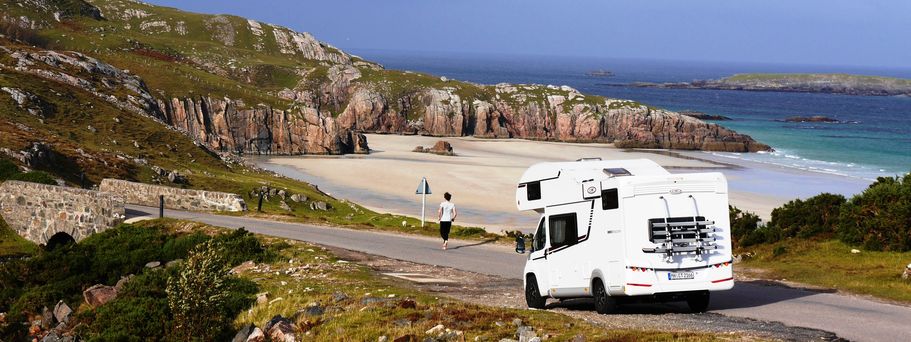 Wohnmobil steht an der Küste auf einem Weg im Hintergrund das Meer und Felsen in England. Ein Mann läuft Richtung Strand. 