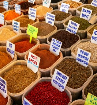 Nahaufnahme von verschiedenen gemahlenen Gewürzen in großen Behältern auf einem Markt, mit englischen und türkischen Beschriftungen.