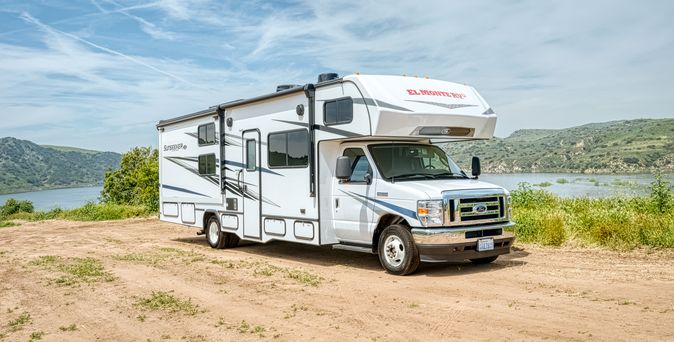  Ein weißes El Monte RV Wohnmobil der Klasse C ist auf einem unbefestigten Platz in der Nähe eines Gewässers und hügeliger Landschaft unter einem leicht bewölkten Himmel geparkt.