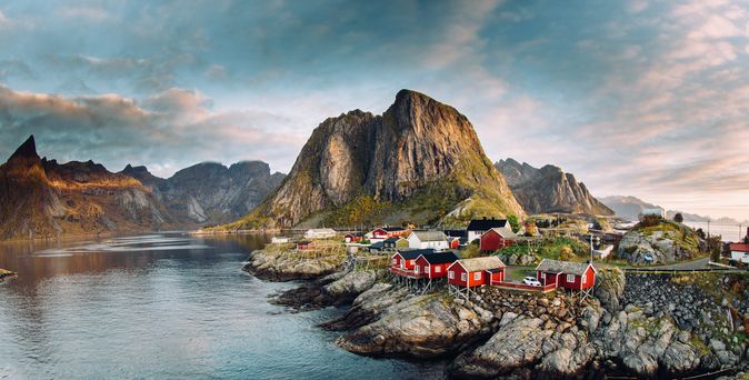 Eine malerische Ansicht der Lofoten-Inseln in Norwegen, die das charmante Fischerdorf Hamnøy mit seinen roten Rorbuer (Fischerhütten) zeigt, umgeben von schroffen Bergen, die sich aus dem Meer erheben.