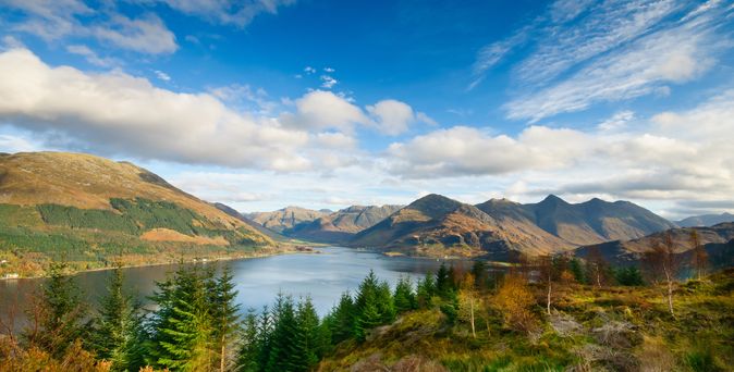 Eine malerische Aussicht auf Loch Ness in Schottland, die den weitläufigen See zeigt, der von bewaldeten Ufern und Bergen umgeben ist, mit einem klaren blauen Himmel darüber.