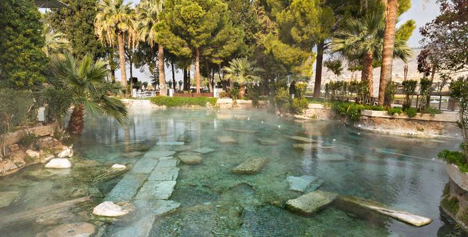  Historisches Thermalbad in Pamukkale, Türkei, mit klarem blauem Wasser und antiken Säulenfragmenten unter Palmen und Bäumen.