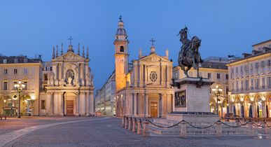 Blick auf den Markplatz Turins in der Dämmerung. Im Vordergrund eine Statur, dahinter ein Brunnen und viele schöne alte Gebäude. Eine Kirche ist ebenfalls im Hintergrund.