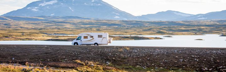 Weißer Campervan fährt auf einem Dammweg, mit Wasser auf der linken und Gras auf der rechten Seite, Berge im Hintergrund.