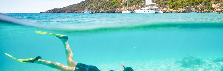 Eine Person schnorchelt in klarem, türkisfarbenem Wasser, während die Sonne die Unterwasserwelt beleuchtet. Felsformationen sind am Rand der Bucht zu sehen.