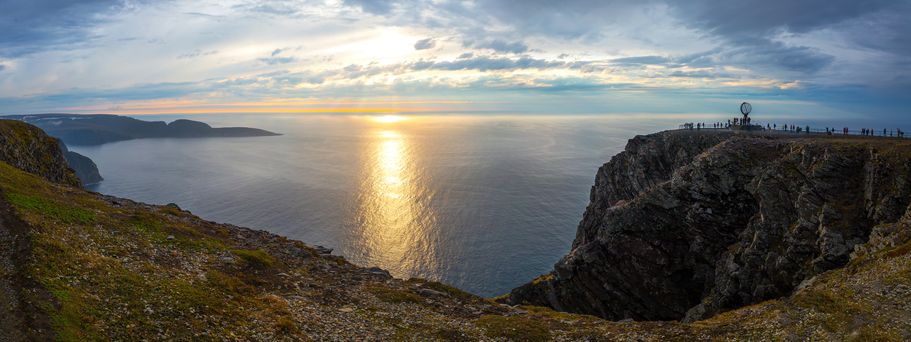 Eine beeindruckende Ansicht des Nordkaps in Norwegen, einem steilen Felsplateau, das ins Arktische Meer abfällt, mit einem Denkmal im Vordergrund und einem weiten, bewölkten Himmel.