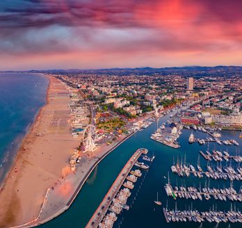 Blick über Rimini im Morgenrot. Auf der linken Seite das Meer, davor der Sandstrand. Rechts davon ein kleiner Hafen und im Hintergrund die Stadt.