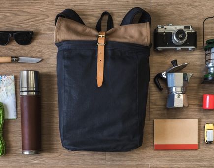 Verschiedene Reiseutensilien wie Rucksack, Sonnenbrille und Kamera liegen auf einem Holztisch.