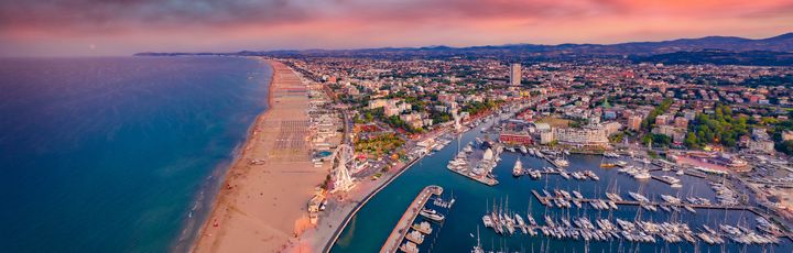 Blick über Rimini im Morgenrot. Auf der linken Seite das Meer, davor der Sandstrand. Rechts davon ein kleiner Hafen und im Hintergrund die Stadt.
