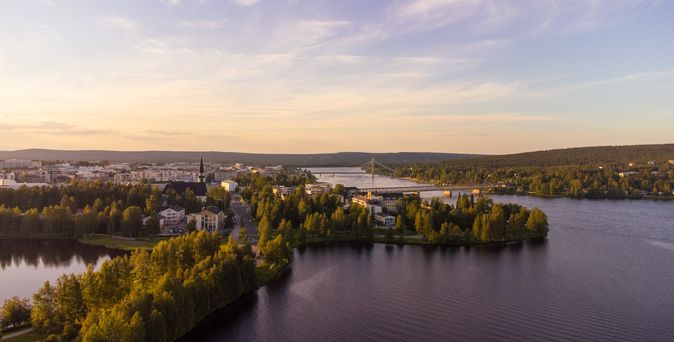 Abendliche Luftaufnahme der Stadt Rovaniemi in Finnland mit Fluss, Brücke und umgebenem Waldgebiet unter sanftem Sonnenlicht. Der Himmel ist leicht bewölkt und in warmen Farben gefärbt.