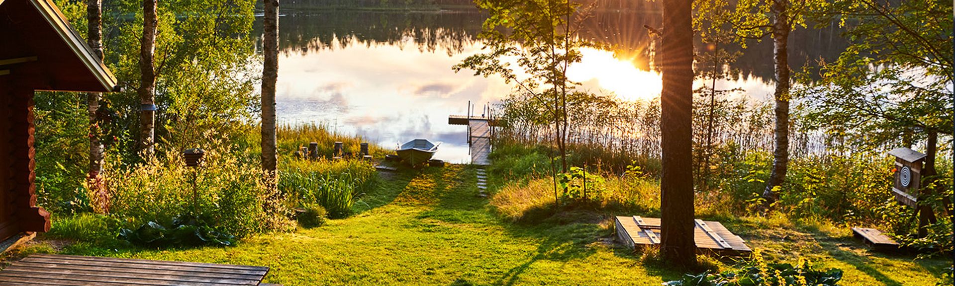 Sonnenuntergang an einem See mit Steg, umgeben von grüner Wiese und Bäumen.