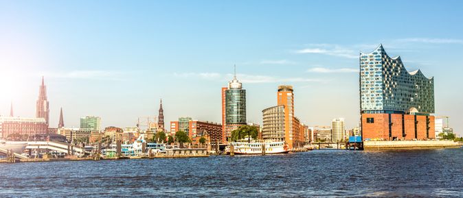 Panoramaaufnahme der Hamburger Skyline mit Elbphilharmonie, Kirchtürmen und modernen Hochhäusern am Ufer der Elbe bei sonnigem Wetter.