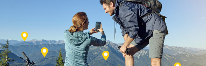 Reisepaar mit Mountainbikes genießt den Panoramablick von einem alpinen Gipfel und hält den Moment mit dem Smartphone fest.