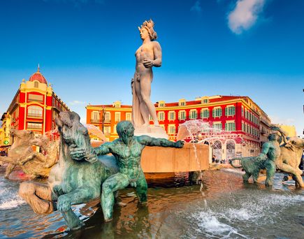 Blick auf den Markplatz, rotes großes Gebäude im Hintergrund. Im Vordergrund einige Statuen in Personenform. Blauer Himmel