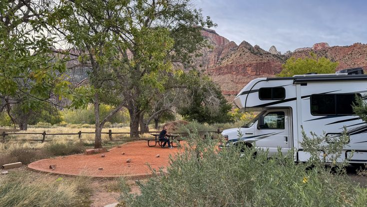 Ein weißes Wohnmobil ist auf einem sandigen Campingplatz unter Laubbäumen geparkt. Im Vordergrund befindet sich ein Picknicktisch mit einer Person, die daran sitzt. Im Hintergrund erheben sich rötliche Felsformationen unter einem leicht bewölkten Himmel.