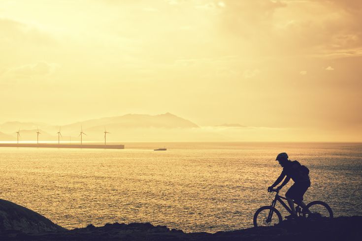 Eine Person fährt mit dem Fahrrad auf einer Straße, die am Meer entlangführt, während die untergehende Sonne den Himmel in warme Farben taucht. Die Szene wirkt ruhig und idyllisch.