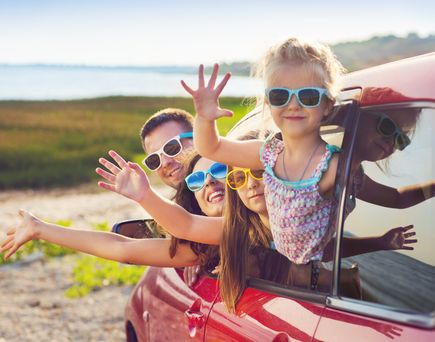 Zwei fröhliche Kinder mit Sonnenbrillen lehnen aus dem Fenster eines roten Autos und winken.