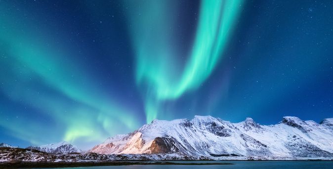 Nordlichter in Finnland über schneebedeckten Bergen und einem Gewässer unter einem klaren Nachthimmel.