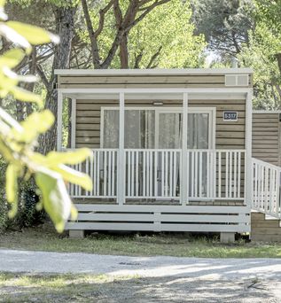 Modernes Mobilheim mit überdachter Veranda im schattigen Pinienwald auf einem Campingplatz.