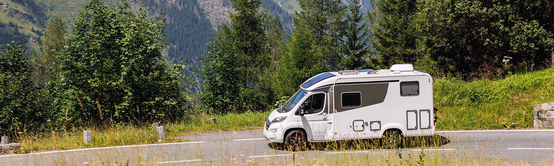 Ein weißes, teilintegriertes Wohnmobil fährt auf einer asphaltierten Landstraße durch eine grüne, hügelige Landschaft.