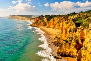 Felsküste in der Algarve in Portugal mit hellblauem Meer, gelb-bräunlichen Felsen und hellbraunem Sand