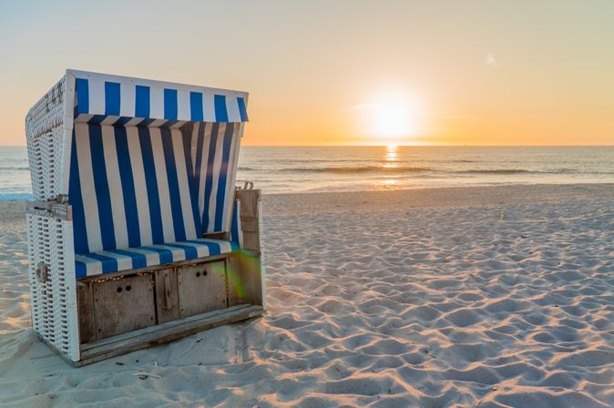 Ein Strandkorb steht am Sandstrand, die untergehende Sonne taucht den Himmel in warme Farben und spiegelt sich auf dem ruhigen Meer.
