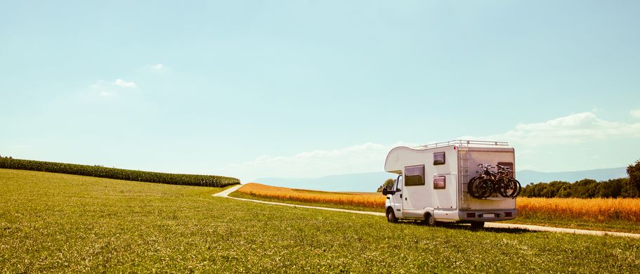 Ein weißes Wohnmobil fährt an einem sonnigen Tag auf einem Feldweg durch weite, grüne und goldene Felder in Richtung Horizont.