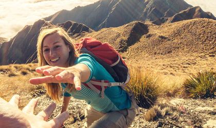 Eine Frau mit Rucksack streckt beim Wandern in den Bergen lachend die Hand nach vorne aus, als würde sie Hilfe beim Aufstieg annehmen.