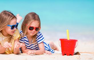 Zwei kleine Mädchen mit Sonnenbrillen spielen am Sandstrand mit bunten Eimern vor türkisblauem Meer.