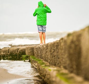 Ein Kind mit grüner Jacke und kurzer Hose spaziert auf einem Steinsteg am Strand.