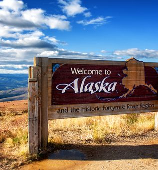 Ein hölzernes "Welcome to Alaska"-Schild mit einer stilisierten Karte von Alaska steht auf einer erhöhten, grasbewachsenen Ebene unter einem weiten blauen Himmel mit weißen Wolken. Im Hintergrund erstrecken sich Berge und Täler.