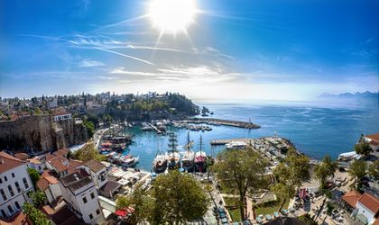 Panoramablick auf den historischen Hafen von Antalya, Türkei, mit traditionellen Segelschiffen, der Altstadt und dem glitzernden Mittelmeer unter strahlender Sonne.