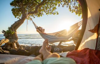 Eine Person liegt entspannt in einer Hängematte unter Bäumen am Strand, Blick auf den Sonnenuntergang über dem Meer.