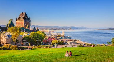 Stadtansicht der Altstadt von Quebec mit Gebäuden, grünem Gras vor dem Sankt-Lorenz-Strom und blauem Himmel an einem Sommerabend. Ein Pärchen sitz auf der grünen Wiese, mit Blick auf Gebäude und Fluss. Blauer Himmel.
